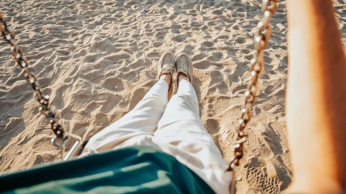 Blick aus einer Strandschaukel auf die eigenen Beine und den Sand
