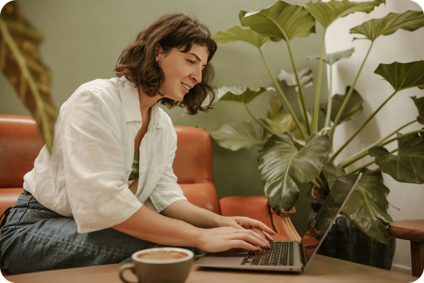 Eine Frau arbeitet sitzend am Laptop, umgeben von Zimmerpflanzen. Nebenbei trinkt sie einen Kaffee.